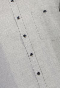Camicia button-up grigia chiara con taschino sul petto, chiuso da un piccolo bottone scuro sulla tasca e sul pannello frontale.
