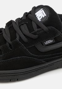 Crni nubuk tenisice s teksturiranim vezicama, crnim kožnim detaljima i istaknutom bijelom Vans logotipu na kružnoj oznaci. Ravni gumeni potplat.