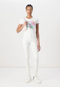 T-shirt blanc avec un imprimé floral coloré, associé à un jean skinny blanc et des baskets blanches, tenant un petit sac à main rose.