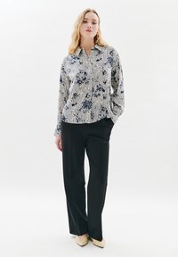 Frau steht da und trägt eine langärmlige gestreifte Bluse mit blauem Blumenmuster, schwarze weit geschnittene Hosen und beigefarbene spitze Schuhe.