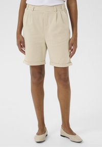 Shorts plissés beiges avec revers roulés, fabriqués en tissu lisse. Comprend une poche avant et une coupe ajustée. Portés avec des ballerines beiges.