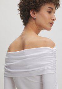 Weißes Off-Shoulder-Oberteil aus geripptem Stoff mit gerafften Details am Ausschnitt, taillierten Ärmeln und glatter Textur.
