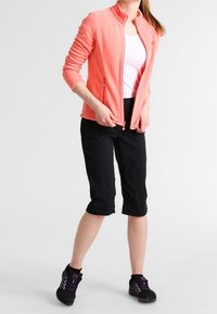 Růžový korálový fleece zip-up blazer přes bílý tílko, v kombinaci s černými kraťasy ke kolenům a černými sportovními botami s fialovými detaily.