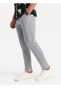 Šedé pletené jogger kalhoty s tapered střihem, s bočními švy, nošeny s bílými teniskami, na prostém světlém pozadí.