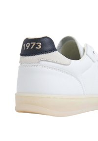 Valkoiset tennarit, joissa on sileä nahkapinta, tummansininen kantatappi, jossa on kohokuvioitu "1973" kultaisella, ja vaalean värinen kumipohja.