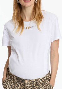 T-shirt en coton blanc à manches courtes, avec un design en écriture dorée "love & all" sur la poitrine. Associé à un pantalon à imprimé léopard.