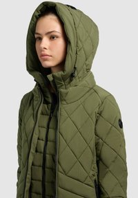 Tűzött olajzöld pufi dzseki magas gallérral, nagy kapucnival és kontrasztos fekete cipzárokkal. Strukturált anyag, átlós varrási részletekkel.