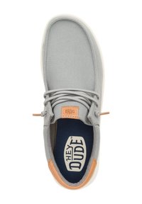 Zapato de lona gris con forma redondeada, que presenta una suela de goma neutra, detalles en marrón, cordones elásticos y una plantilla azul marino con el logo.