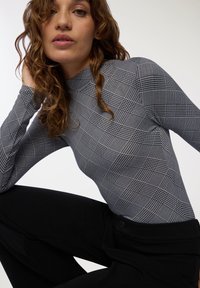 Giovane donna con capelli ricci che indossa una camicia a maniche lunghe a quadri bianco e neri e pantaloni neri, in posa su uno sfondo semplice.