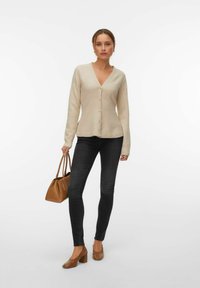 Femme portant un cardigan beige boutonné, un jean skinny noir, et des chaussures à talons carrés marron, tenant un sac à main marron sur fond blanc.
