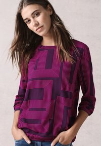Blusa viola con linee geometriche rosa, maniche lunghe con polsini arricciati, scollatura rotonda e vestibilità leggermente ampia. Indossata con jeans casual.
