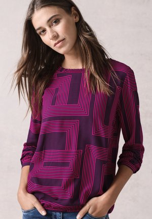Blusa viola con linee geometriche rosa, maniche lunghe con polsini arricciati, scollatura rotonda e vestibilità leggermente ampia. Indossata con jeans casual.
