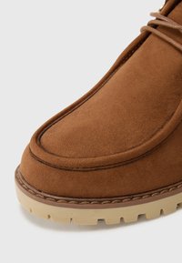 Botte en daim marron avec un bout arrondi, des coutures contrastantes autour du bord et une semelle en caoutchouc beige clair texturée.