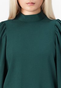 Tyrkysová halenka s dlouhými rukávy, vysokým výstřihem, strukturovanou tkaninou a nařasenými detaily na ramenech. Hladký, volný silueta.