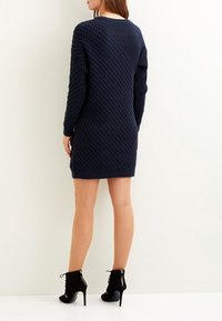 Abito maglione lavorato a maglia di colore blu navy con un motivo a diamante testurizzato e maniche lunghe, dotato di orlo dritto e indossato con stivali neri alla caviglia.