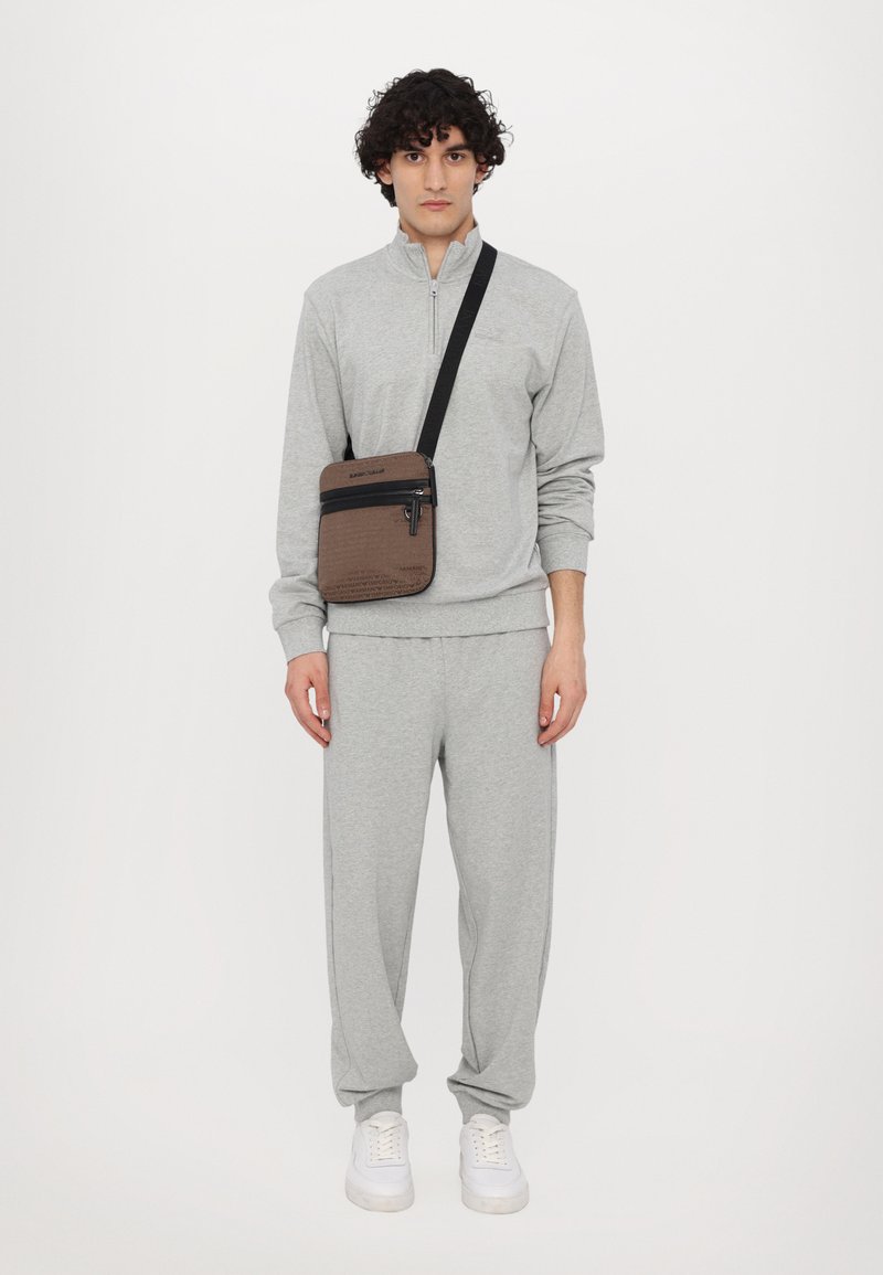 Pull-over de sport gris avec demi-fermeture éclair, pantalon de survêtement gris et sac bandoulière marron avec deux poches zippées. Des baskets blanches complètent le look.