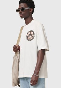 Hvid oversized t-shirt med cirkulært fredssymbol grafisk i jordfarver, korte ærmer og en afslappet pasform; parret med grå solbriller og en beige taske.
