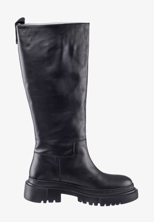 Femme portant un gilet en cuir noir sans manches surdimensionné avec ceinture, un haut noir à manches longues, un pantalon en cuir et des bottes noires montant jusqu'aux genoux sur fond blanc.