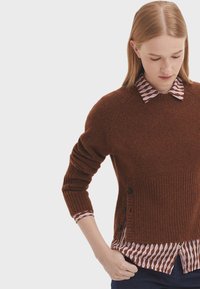 Brauner Wollpullover mit geripptem Saum und Seitenknöpfen, über einem Hemd mit einem geometrischen Muster in Pink und Creme geschichtet.