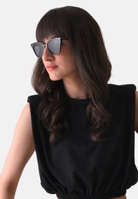 Černé sluneční brýle ve tvaru kočičích očí se šedými skly, kovovými detaily a elegantním designem. Modelka má na sobě černý top bez rukávů se skládáním v pase.