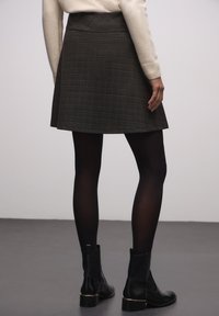 Harmaa ruudullinen liehuva hame, jossa on sileä vyötärökaiha, yhdistettynä mustiin sukkahousuihin ja nilkkureihin, takaviistosta nähtynä.