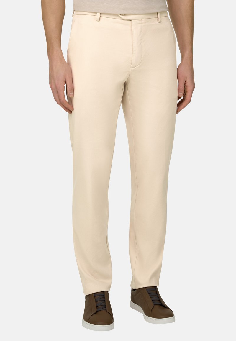 Homme portant un pantalon beige slim et des baskets marron foncé, debout devant un fond clair uni.