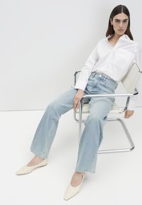 Frau mit langen dunklen Haaren, die ein locker sitzendes weißes Hemd und hellblaue Jeans mit weitem Bein trägt, sitzt auf einem modernen Stuhl aus Chrom und Weiß.