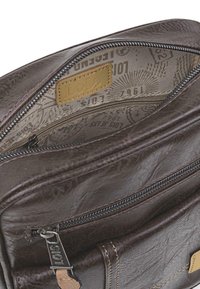 Interior de una bolsa de cuero marrón con un forro de tela estampada. Cuenta con un bolsillo con cremallera, acentos dorados y una etiqueta de logo visible.