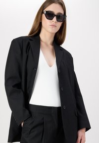 Svart oversized blazer med klaffkrage, enkelt lomme og knappelukking, brukt over en hvit V-hals topp og svarte bukser. Svarte solbriller.