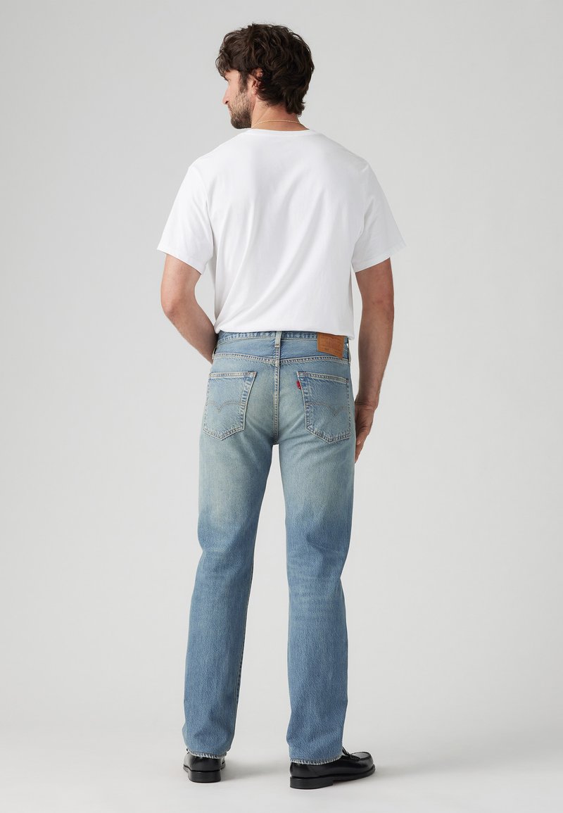 Jean en denim bleu clair avec une coupe décontractée, comportant des poches arrière et un patch en cuir beige. Associé à une chemise blanche à manches courtes.