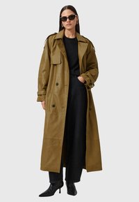 Olijfgroene leren trenchcoat met een oversized ontwerp, voorzien van een kraag, grote zakken en vijf knopen; gecombineerd met een zwarte top en hoge laarzen.