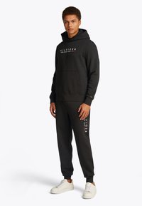 Black sweatshirt with hood and front pocket, featuring a logo. Paired with matching tapered sweatpants and white sneakers.