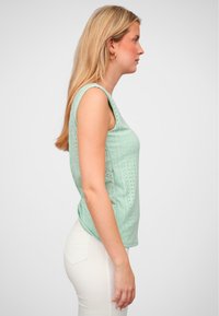 Top verde deschis fără mâneci, realizat dintr-un material texturat cu mici perforații. Guler rotund și o croială lejeră, asortat cu pantaloni albi.