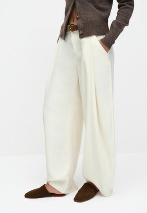 Weite weiße Hose, getragen mit einem braunen Gürtel, braunem Cardigan und passenden braunen Slip-On-Schuhen auf hellem Hintergrund.