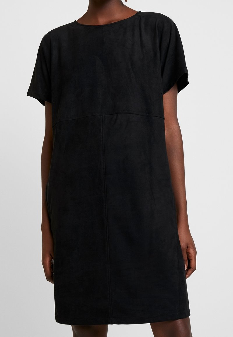 Femme portant une robe noire à manches courtes, ample, avec un col rond et des détails de coutures visibles à l'avant.