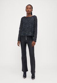 Zwarte lange mouwen top met een textuur en geborduurd patroon. Gecombineerd met strakke zwarte broek met knopen aan de voorkant. Draagt zwarte hoge hakken.