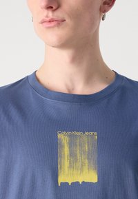 T-shirt de algodão azul com um design gráfico amarelo apresentando linhas verticais e o texto "Calvin Klein Jeans" centralizado no peito. Mangas curtas.