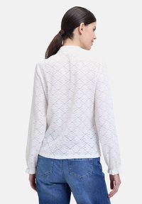 Blusa bianca con tessuto testurizzato e motivo in pizzo, maniche lunghe e una leggera lucentezza; abbinata a jeans blu, con una vestibilità comoda.