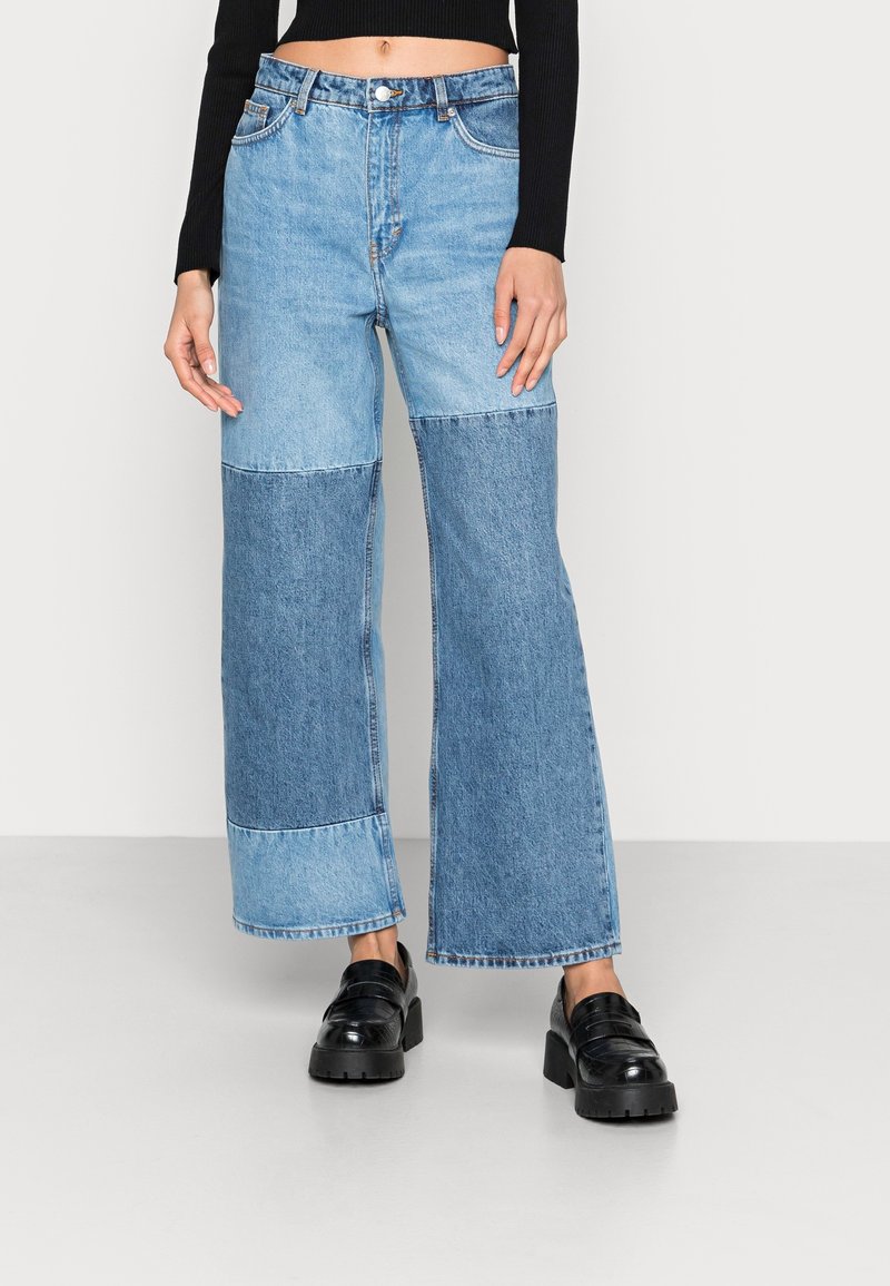 Femme portant un jean large bicolore bleu, un haut noir court à manches longues et des mocassins noirs à plateforme épaisse, debout sur un sol gris.