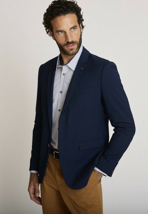 Homme portant un blazer bleu marine sur une chemise à motifs boutonnée et un pantalon marron, posant avec une main dans la poche, regardant sur le côté.