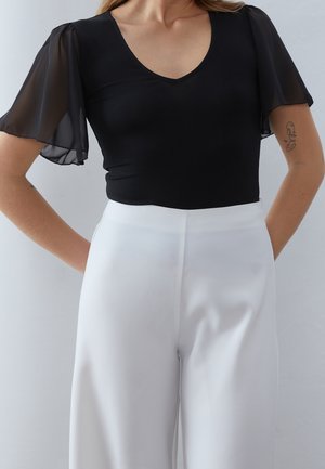 Top nero aderente con maniche trasparenti corte e scollatura a V, abbinato a pantaloni bianchi a gamba larga a vita alta. Tessuto liscio e linee pulite.
