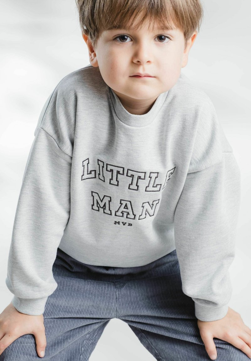 Jeune garçon aux cheveux bruns portant un sweat gris marqué « Little Man » et un pantalon en velours côtelé bleu, penché en avant avec les mains sur les genoux.