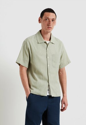 Giovane uomo con lentiggini che indossa una camicia verde chiaro a maniche corte sopra una t-shirt bianca e pantaloni blu scuro, in piedi con una mano in tasca.