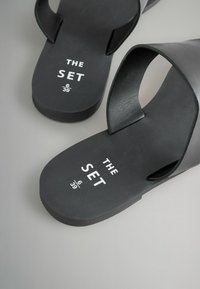 Mustad mustad sandaalid, millel on sujuv, matt viimistlus ja laiad rihmad. Kaunistatud valge teksti "THE SET" ja suuruse detailidega.