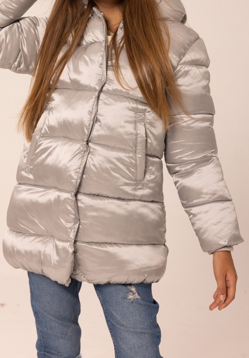 Personne portant une doudoune brillante gris clair, avec de longs cheveux bruns et un jean bleu déchiré, posant avec un bras levé et l'autre détendu vers le bas.