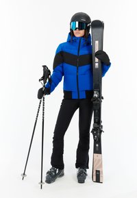 Modrá a čierna puffa bunda, čierne lyžiarske nohavice, rukavice, drží lyže a palice. Čierna prilba s zrkadlovými okuliarmi. Šedé lyže.