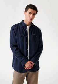 Dunkelblaues Langarmhemd mit Knopfleiste und zwei Brusttaschen, weißen Knöpfen und einer weichen Textur, über einem weißen T-Shirt getragen.