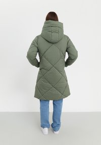 Parka rembourrée en vert olive avec capuche, dotée d'un motif en losanges et d'une coupe mi-longue, associée à un jean bleu clair et des baskets blanches.