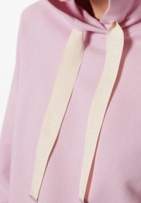 Sudadera rosa con una textura suave, que presenta una capucha amplia y cordones beige con puntas plateadas. Diseño minimalista sin patrones.