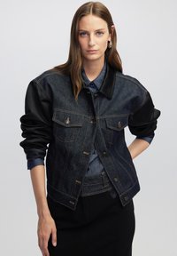 Giubbotto di jeans con maniche in pelle nera, due tasche sul petto, cuciture dorate, chiusura a bottoni e colletto strutturato.